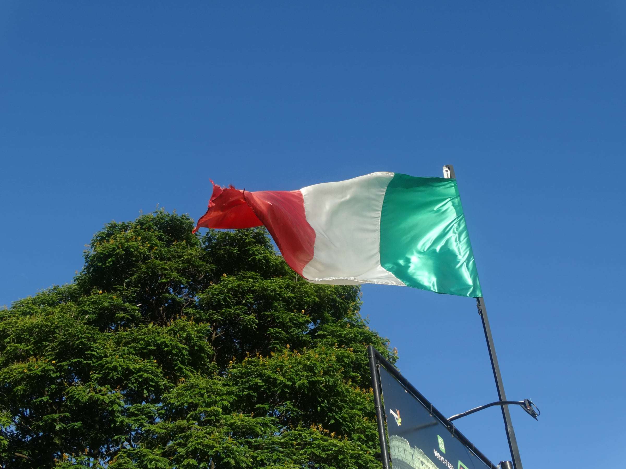 Bandeira da Itália tremulando no mastro contra o céu azul, acima das árvores