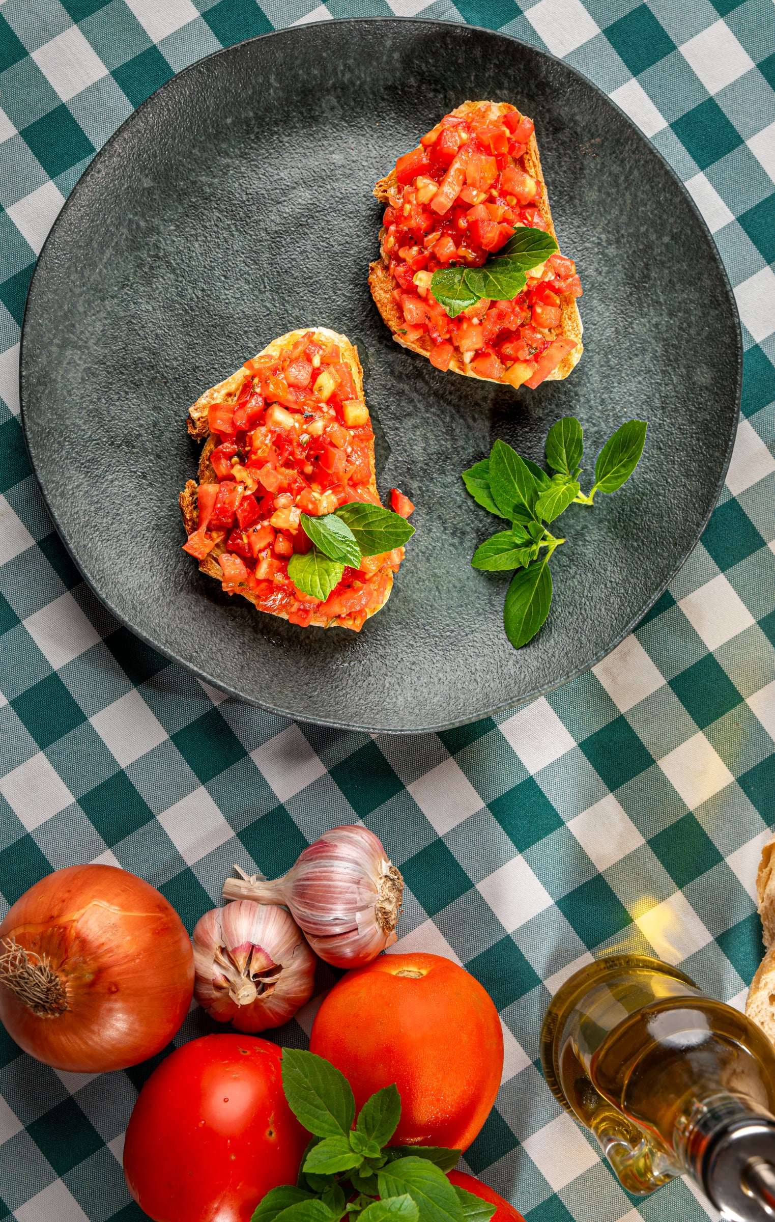 Duas fatias de bruschetta com tomate picado e manjericão em prato cinza, ao lado de tomates, cebola e alho