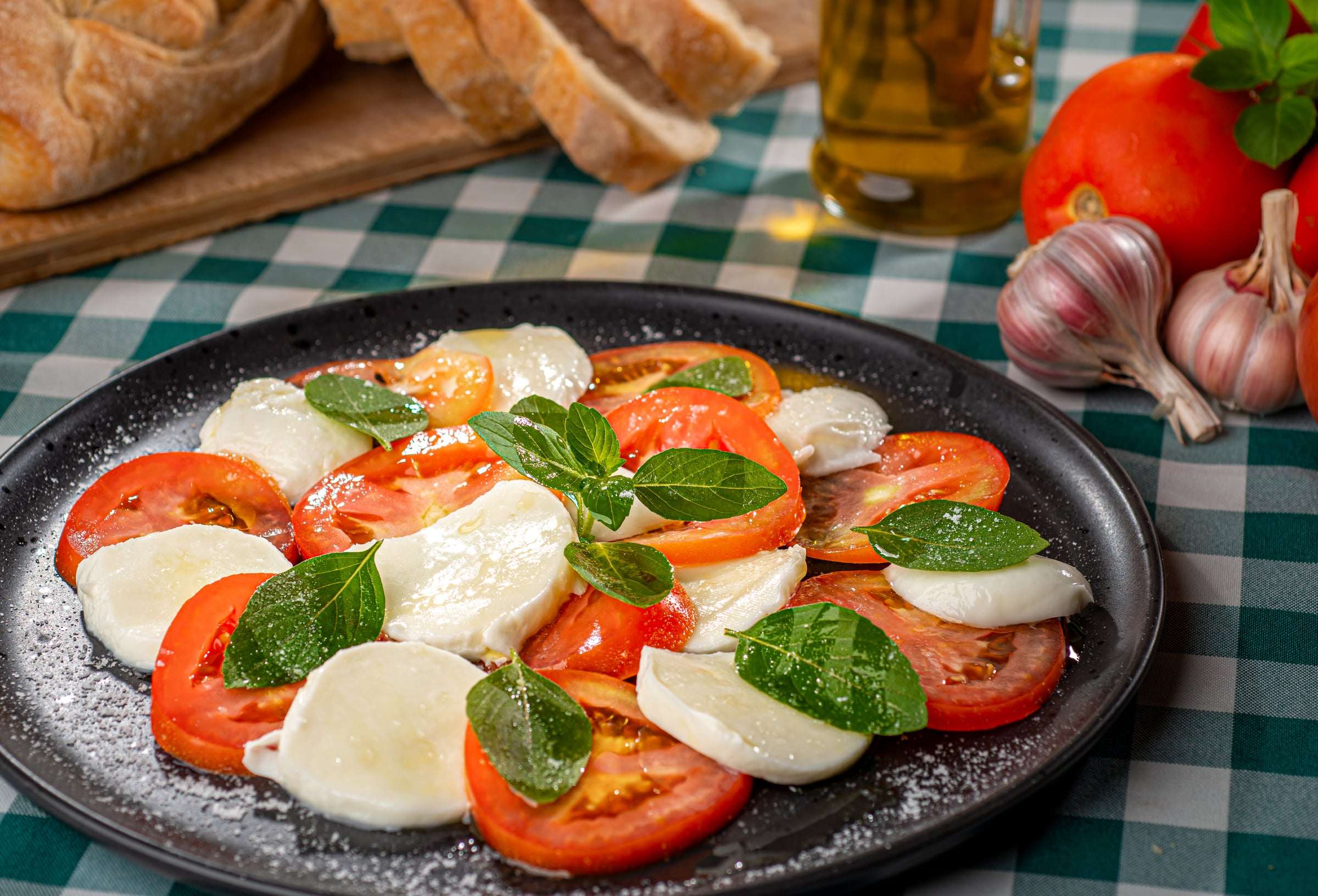 Caprese em prato preto: fatias de tomate e mozzarella de búfala com folhas de manjericão
