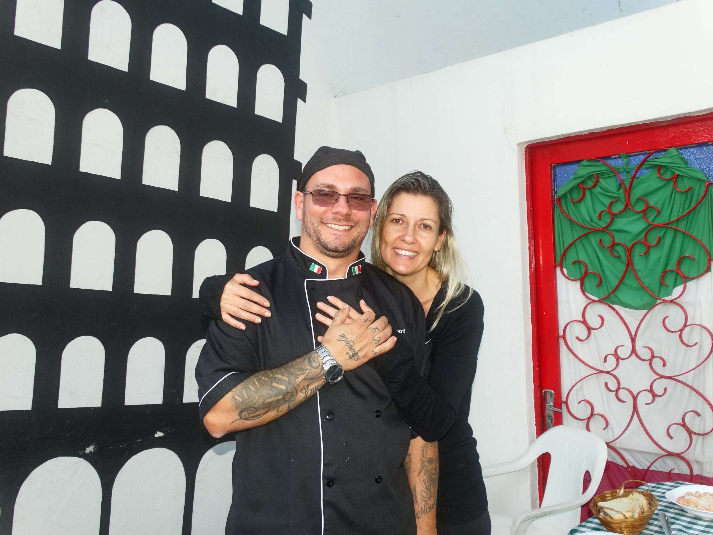 Chef Alessandro Giaccari abraçado a Christiane Gonçalves diante do mural do Coliseu