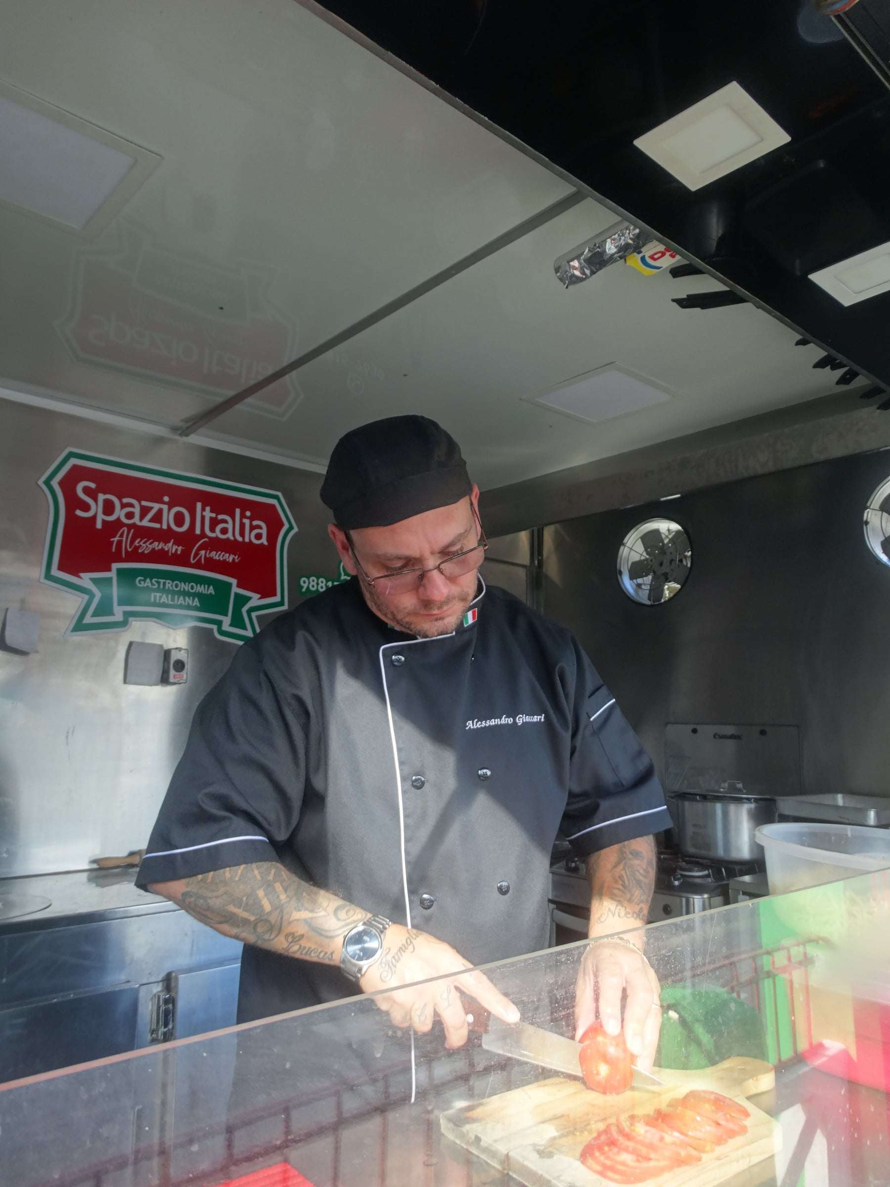 Chef Alessandro Giaccari fatiando tomates na bancada do food truck, com o selo do Spazio Italia na parede
