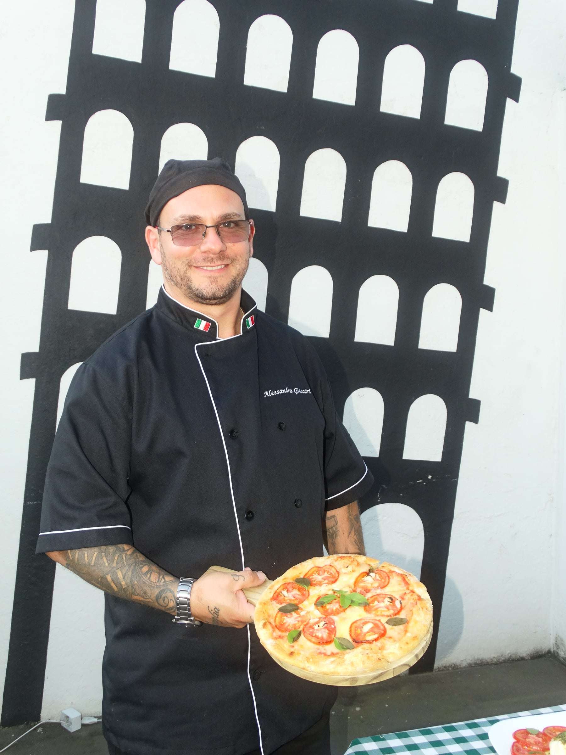 Chef Alessandro Giaccari, de dólmã preto, segurando uma Pizza Margherita diante do mural do Coliseu