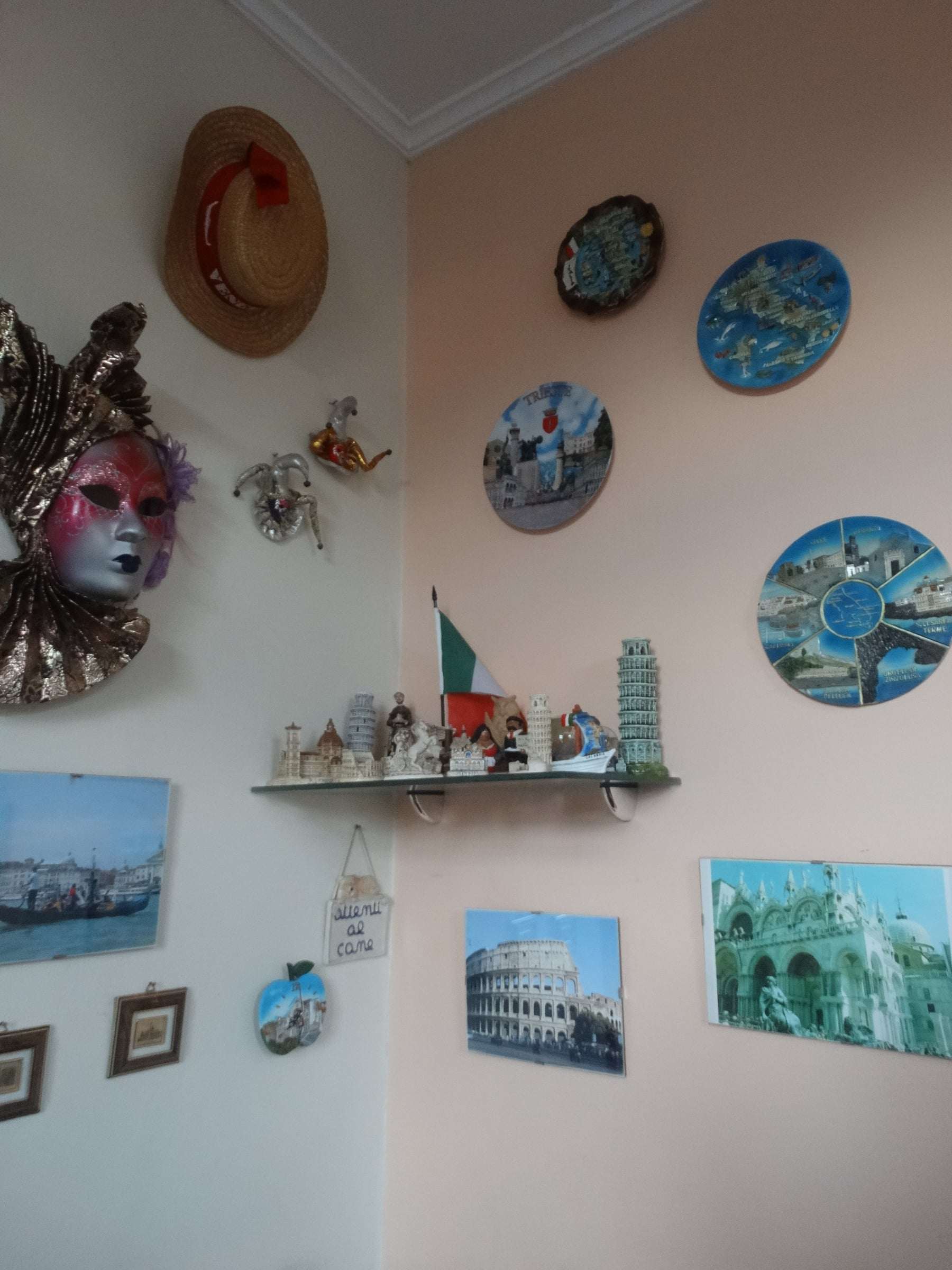Canto do salão com máscara veneziana, chapéu de palha, pratos decorativos, miniaturas e fotos da Itália na parede