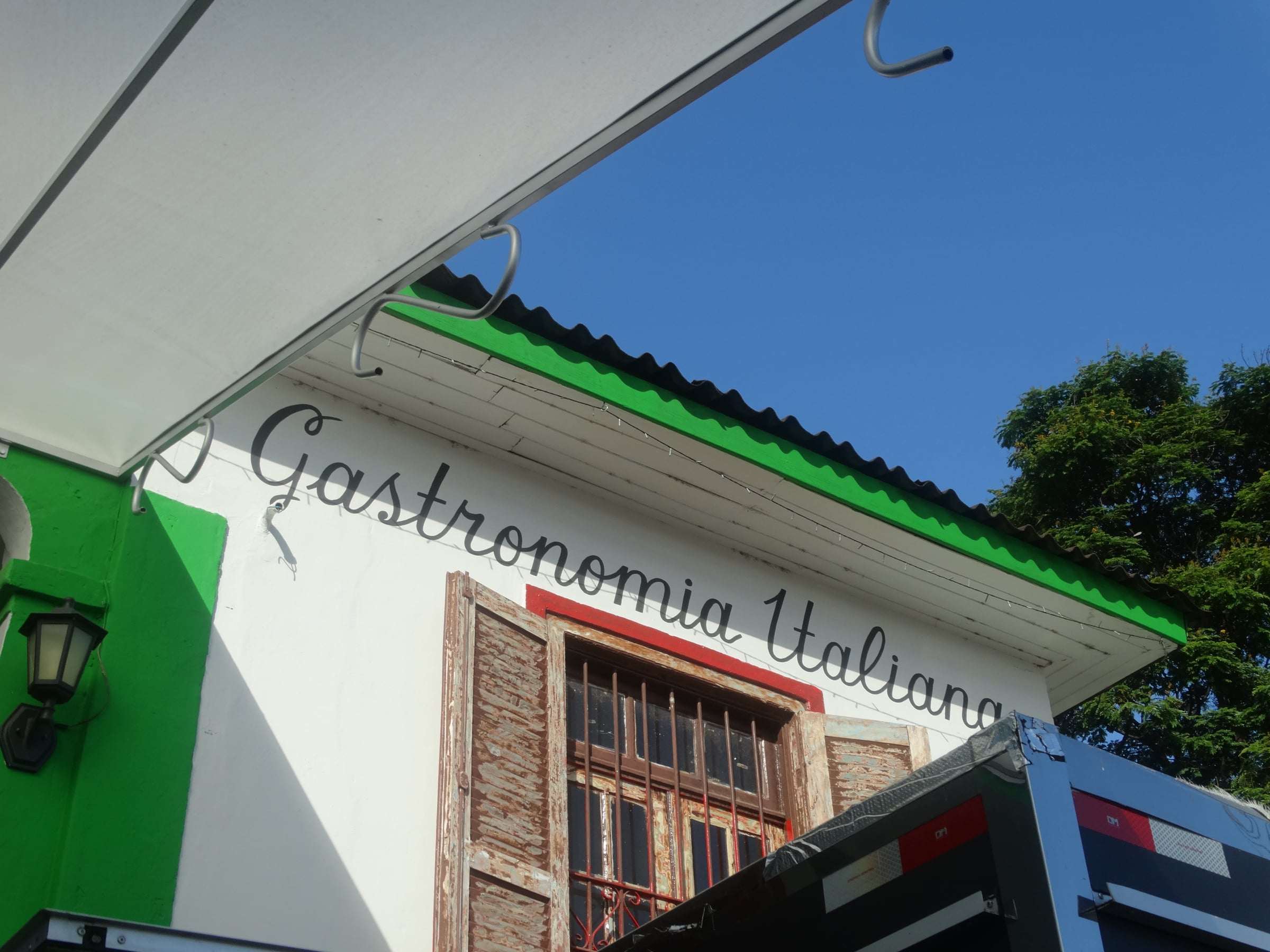 Fachada branca e verde com o letreiro Gastronomia Italiana e a janela de madeira, contra o céu azul