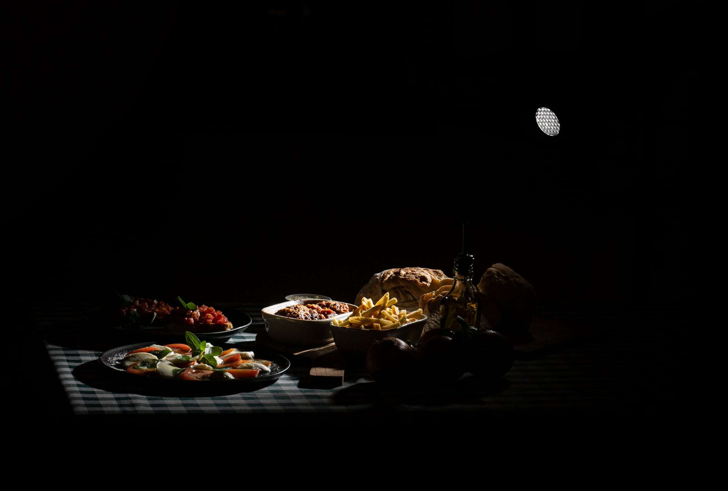 Mesa à meia-luz com bruschetta, caprese, batata frita e pão italiano sobre toalha xadrez