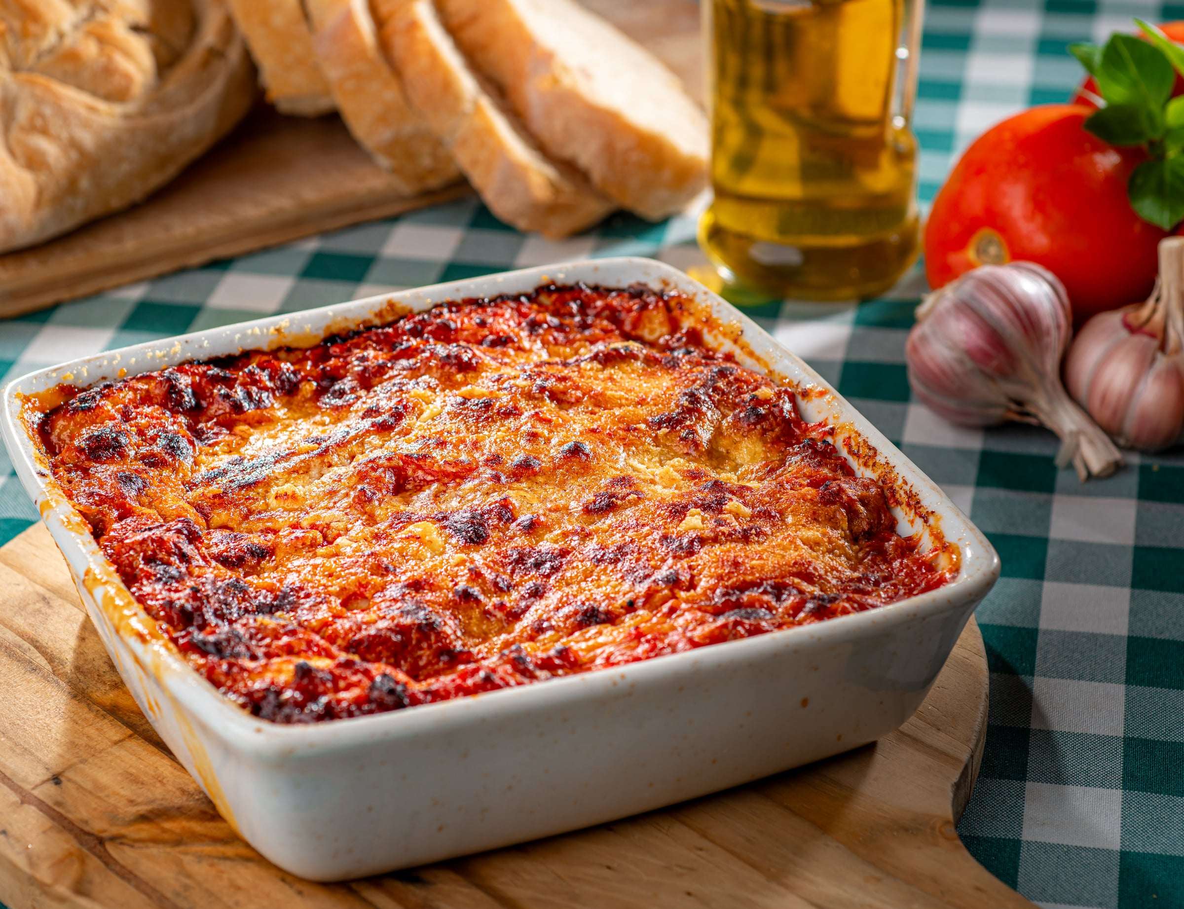 Lasanha gratinada em travessa branca sobre tábua de madeira, com pão italiano e azeite ao fundo