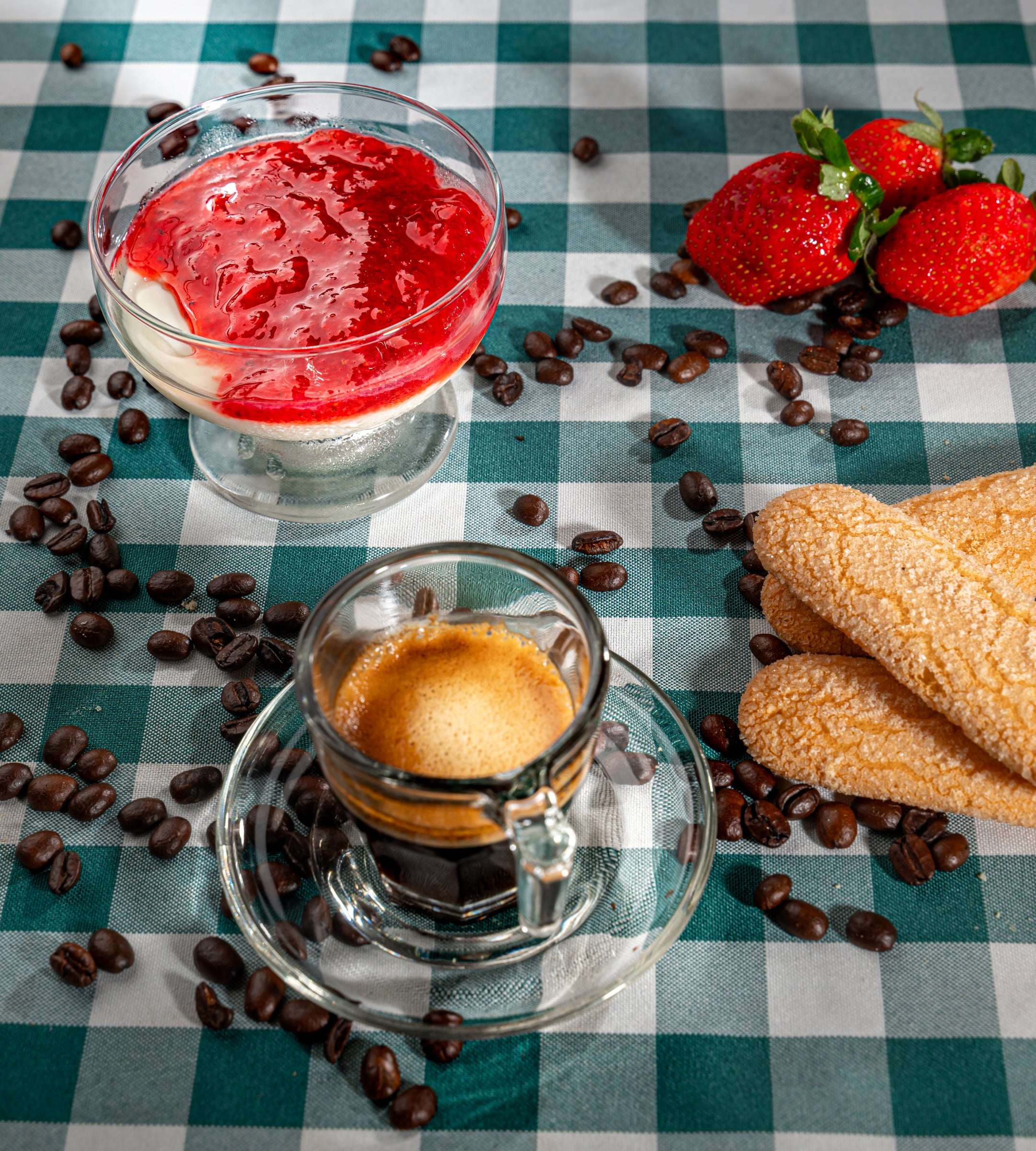 Panna cotta com calda de morango em taça de vidro, ao lado de um café expresso e grãos de café