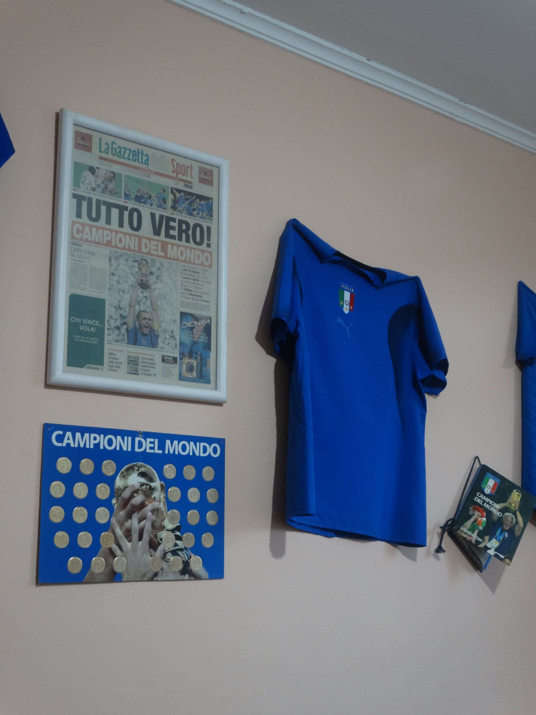 Parede com a capa de jornal italiano Tutto vero!, a camisa azul da seleção italiana e o quadro Campioni del mondo