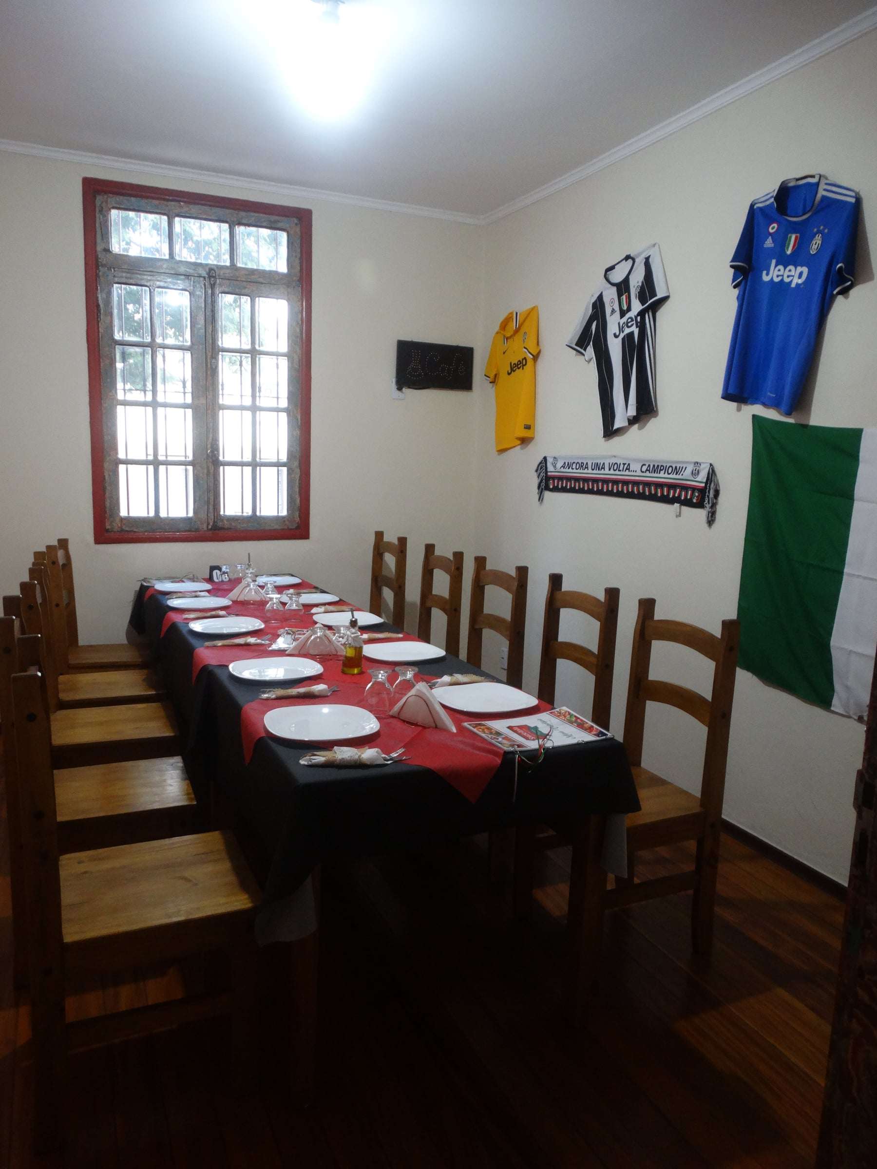 Salão interno com mesa comprida posta, cadeiras de madeira, camisas de futebol italianas e a bandeira da Itália na parede