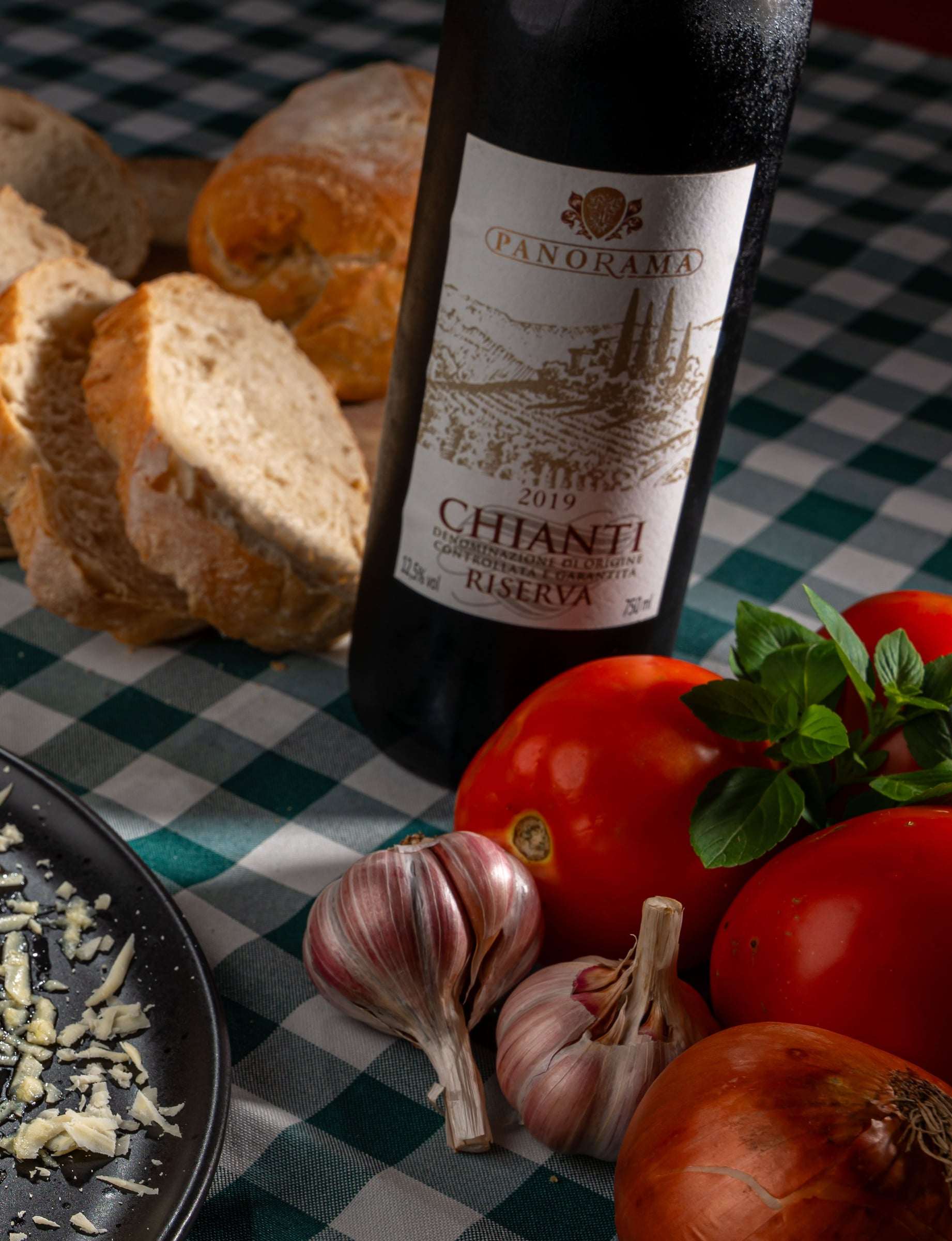 Garrafa de vinho tinto Chianti Riserva na mesa com toalha xadrez, ao lado de pão italiano, tomates, alho e cebola